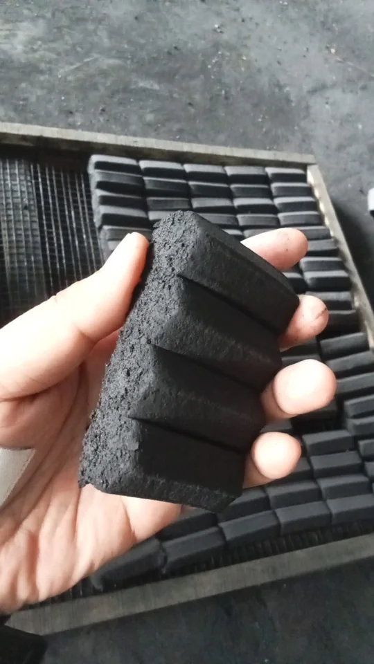 A hand holding a freshly pressed DRAGON briquette bar, scored into cubes, in front of drying racks